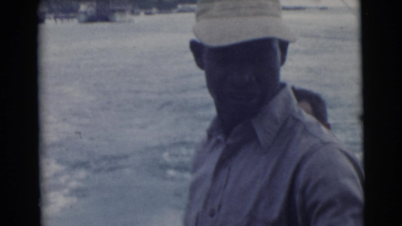 Man in a cap and light shirt stands on a boat with open water behind him.
