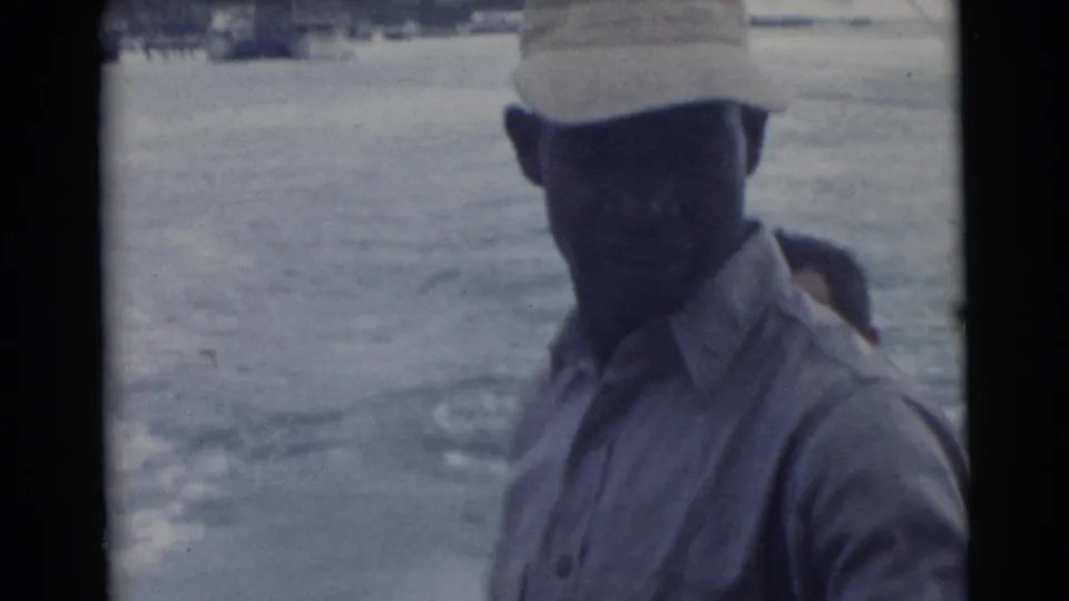 In 1956 Miami home-movie footage, a man in a cap and light shirt stands aboard a boat against a soft blue view of the water.