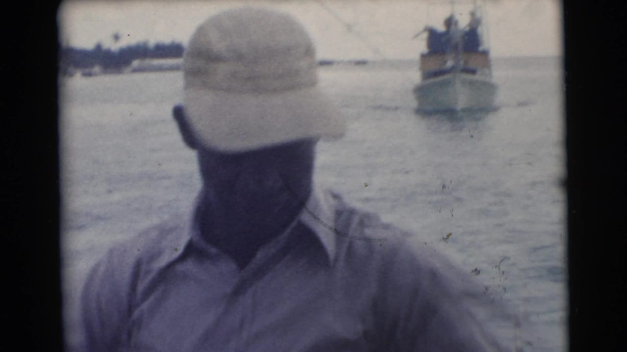 Man in a cap faces downward on a boat while another vessel appears in the distance.