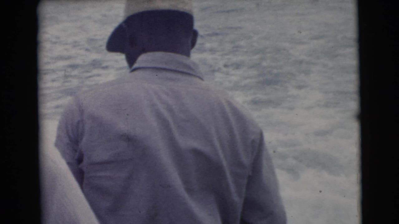 Man in dark cap and light jacket seen from behind beside open water.