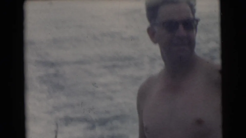 1956 Miami home-movie frame with a shirtless man in sunglasses close to camera, water visible behind him.