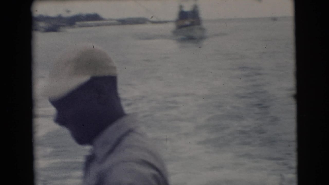 Man in a light cap stands in profile on a boat with open water and distant vessels behind him.