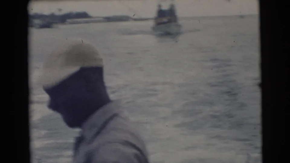 1956 Miami home-movie frame showing a man in a light cap near the edge of a boat, with open water and distant boats in soft focus.