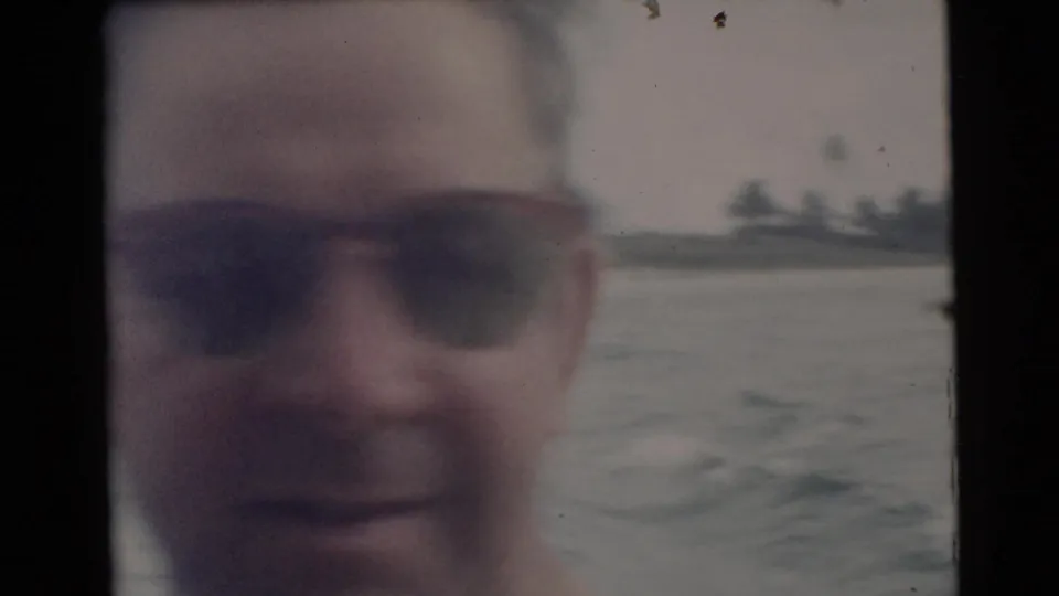 1956 Miami home-movie close-up of an adult man in sunglasses aboard a moving boat, with water and palm-lined shore behind him.