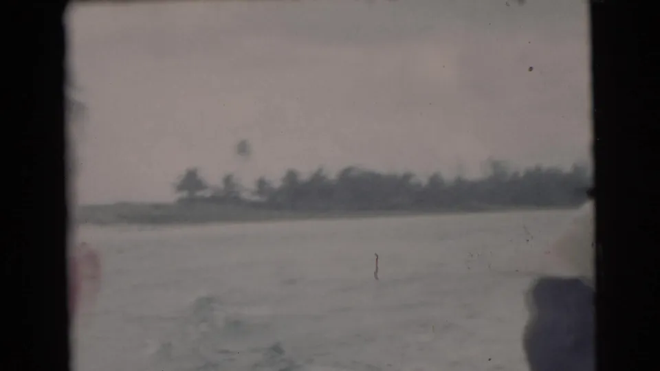 1956 Miami home-movie frame looking from a boat across calm river water toward a low, tree-lined shoreline under an overcast sky.