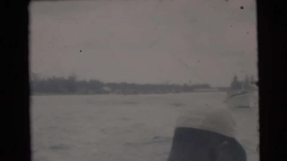 1956 Miami boat-view frame with calm water, distant shoreline, and an adult man partly visible at the right edge.