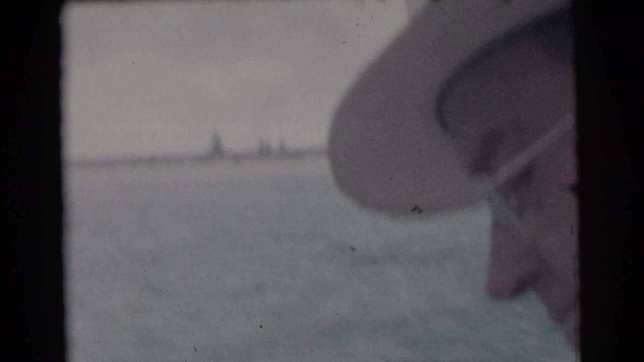 Partial profile of a man in glasses and a hat beside open water and distant shoreline.