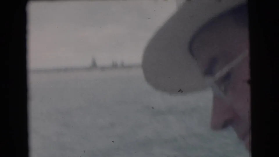 1956 Miami-area 8mm frame with a partial adult face in glasses and a hat near the boat edge, with water and a low shoreline beyond.