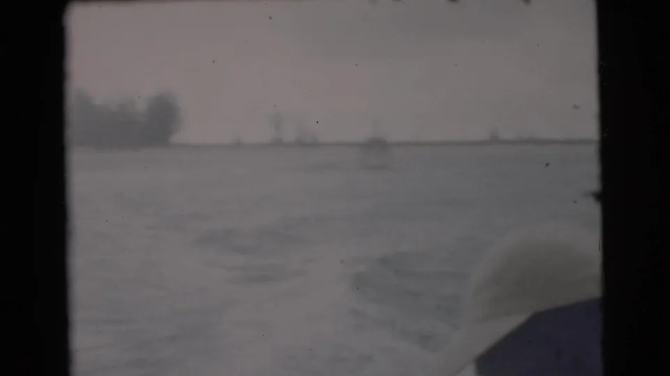 1956 Miami-area 8mm frame looking back from a moving boat across wake, water, and a hazy tree-lined shore.