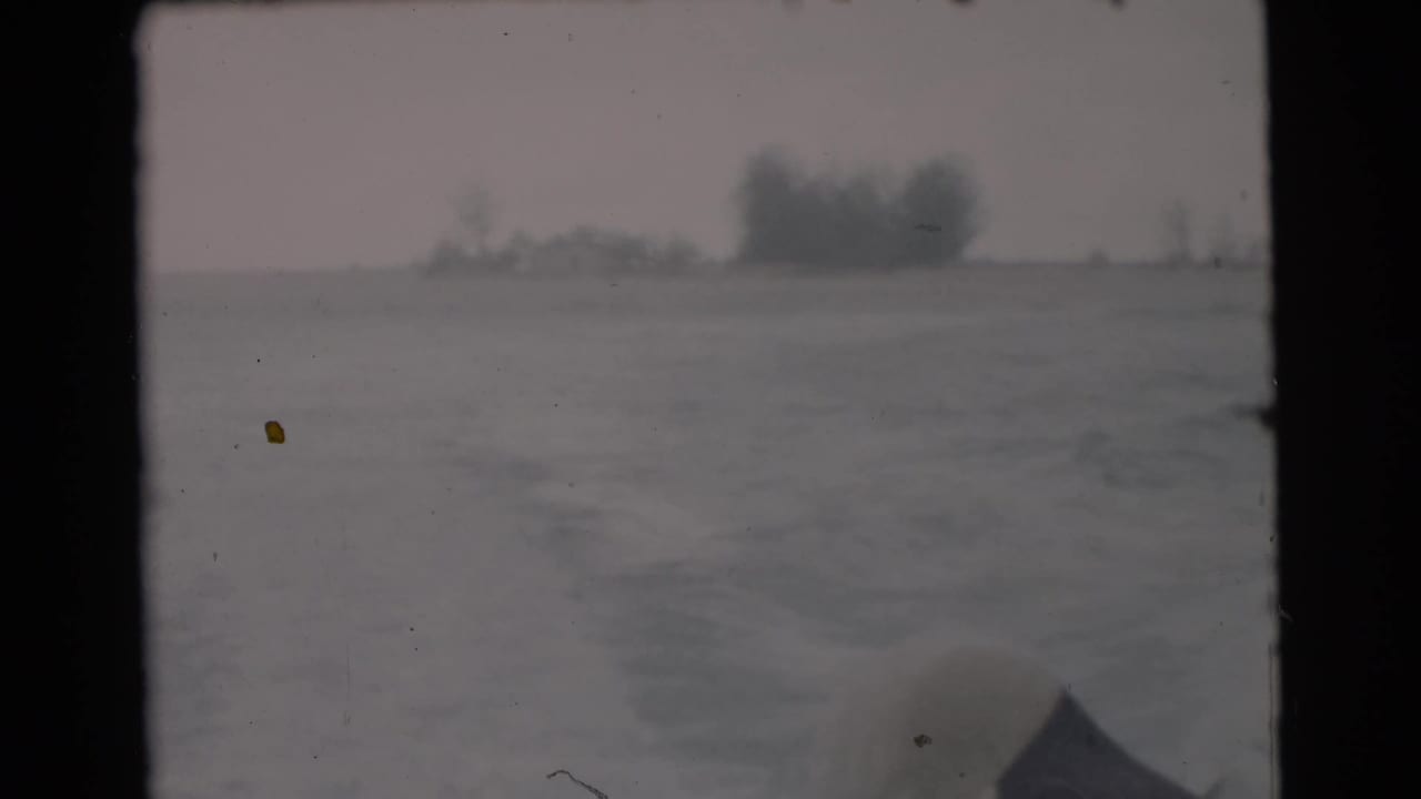 1956 Miami-area home-movie view from a moving boat, showing water, faint wake, shoreline trees, and a dark boat edge.