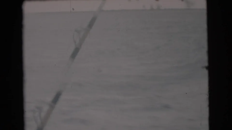 1956 Miami-area boat POV with a fishing rod crossing the frame above rippled water and a distant low shoreline.