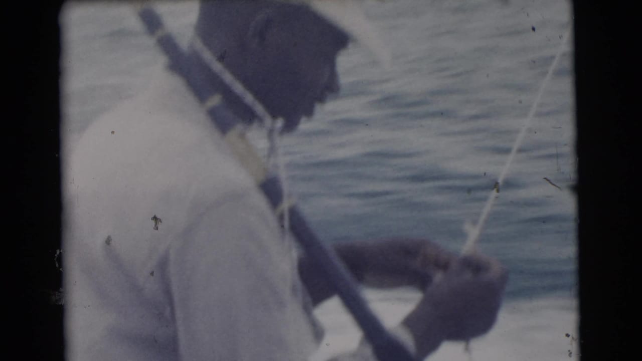 1956 Miami home-movie frame showing a man in a light shirt handling fishing line beside a rod with open water behind him.