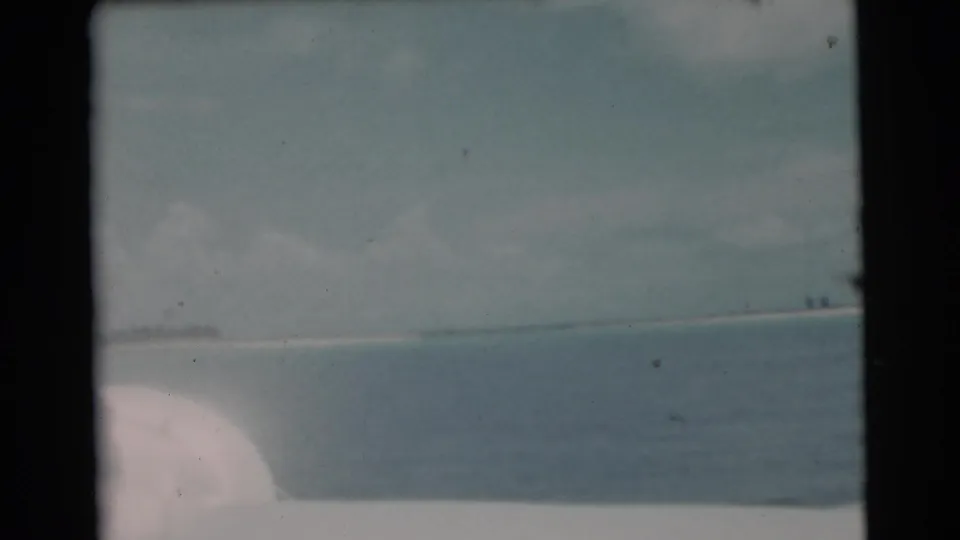 1956 Miami coastal view from the boat, with calm sea, a low shoreline, and the fisherman mostly out of frame.
