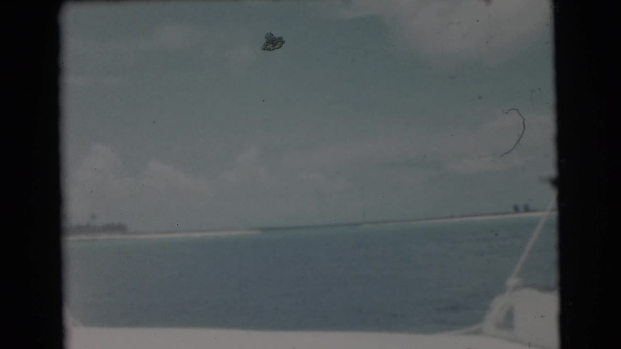 1956 Miami 8mm frame showing calm water, a low shoreline, cloudy sky, and a rod or boat line entering at the right edge.