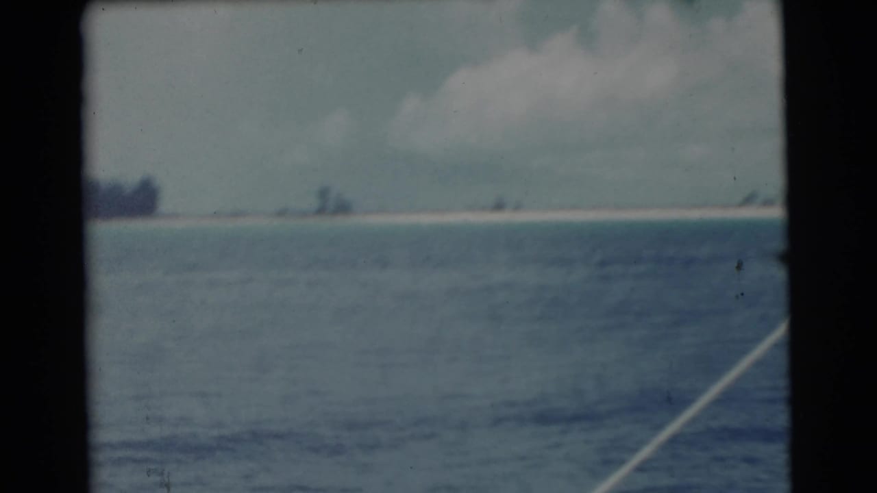 1956 8mm view of open water and a distant Miami-area sandy shoreline through a dark window frame.