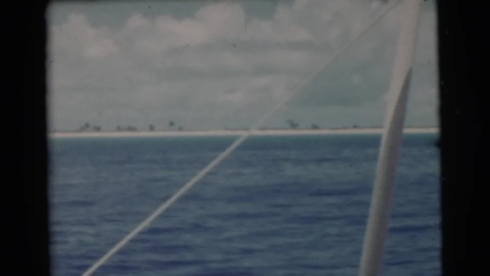 1956 8mm coastal view from a boat, showing ocean, a distant sandy strip, and a pale rail or cable near the right side.
