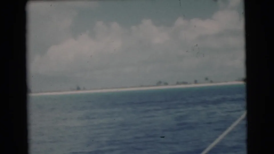 1956 8mm Miami-area ocean view with a tilted horizon, distant sand beach, and small palm silhouettes under a cloudy sky.