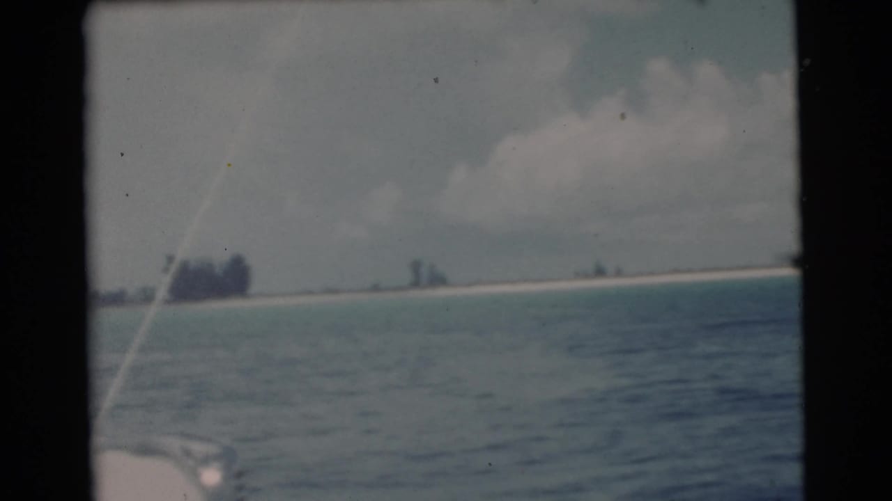 View across blue-green water toward a low island shoreline from a sailboat.