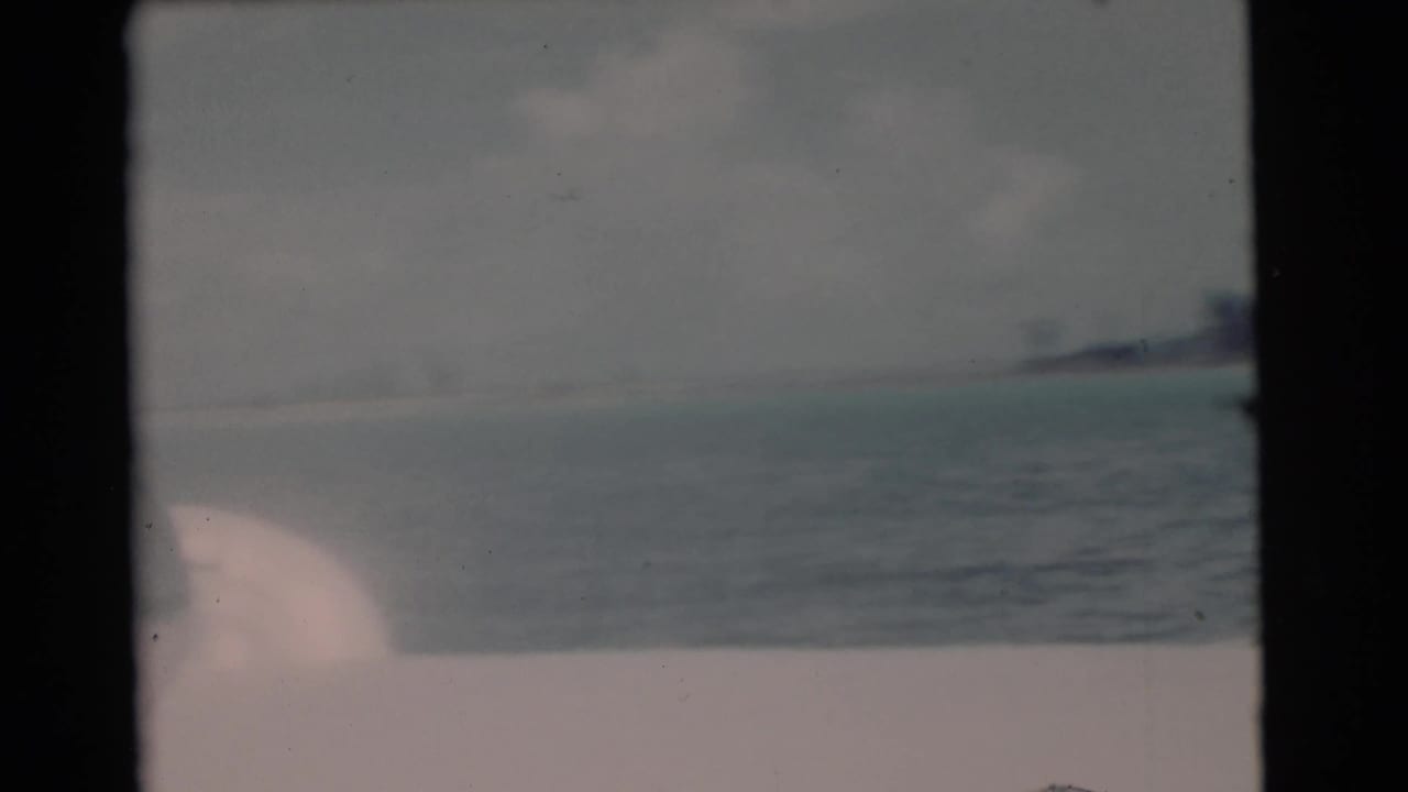 Faded 8mm frame of open coastal water near Miami, with a low shoreline and a pale boat edge or sail shape in the foreground.