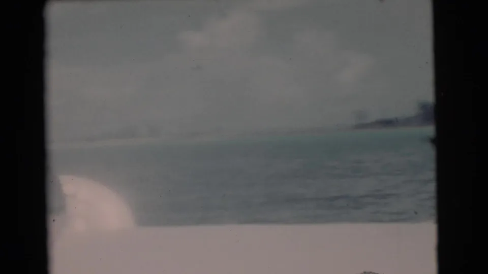Faded 8mm frame of open coastal water near Miami, with a low shoreline and a pale boat edge or sail shape in the foreground.