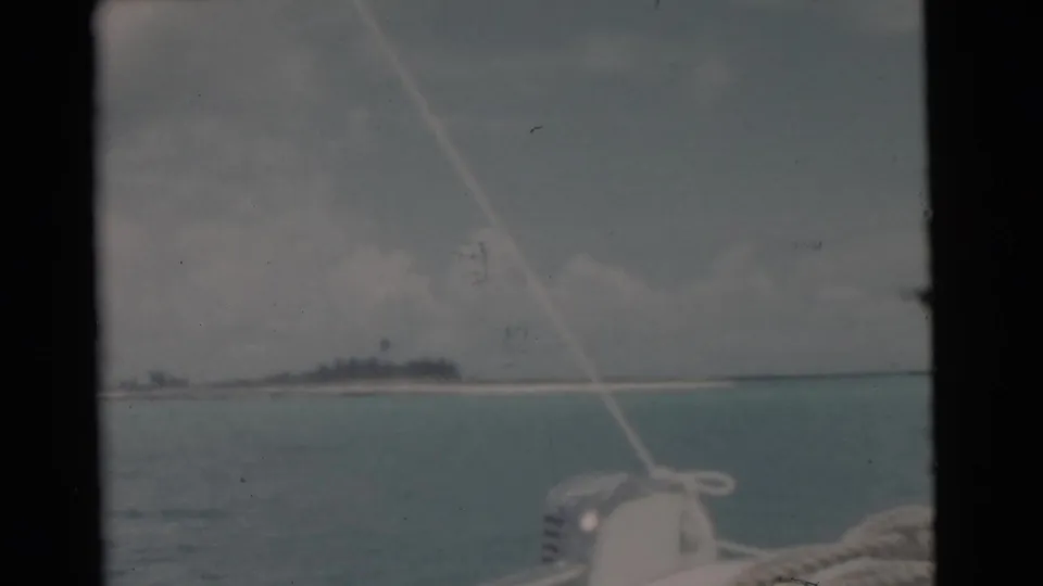 1956 home-movie boat-deck view near Miami, showing sailboat rope and hardware against turquoise water and a low island shoreline.