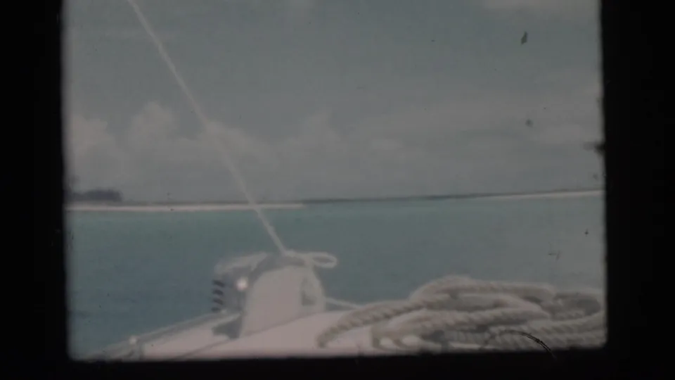1956 Miami-area 8mm frame from a boat deck, with coiled rope in front and blue water leading to a distant low island.