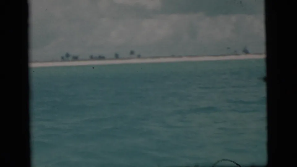 1956 Miami-area 8mm view across blue ocean toward a low sandy island under a cloudy sky.