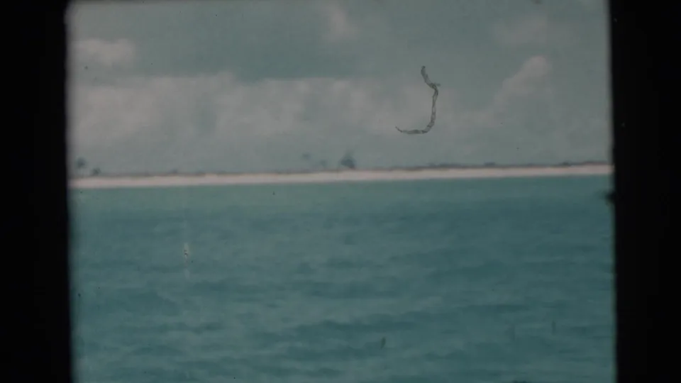 1956 Miami-area 8mm frame showing turquoise water, a white distant beach, and cloudy sky through dark vertical framing.