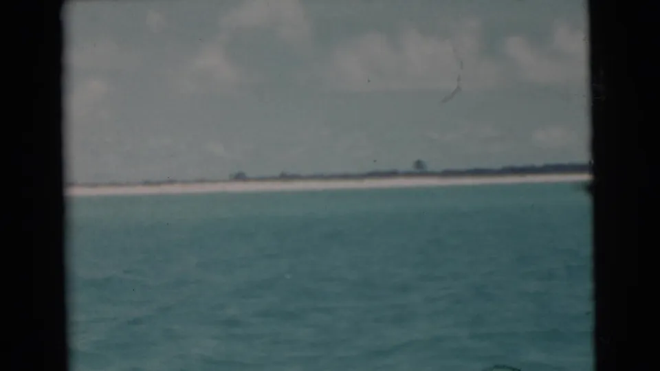 1956 Miami-area 8mm frame with calm turquoise water, a thin beach line, low dark vegetation, and cloudy sky.