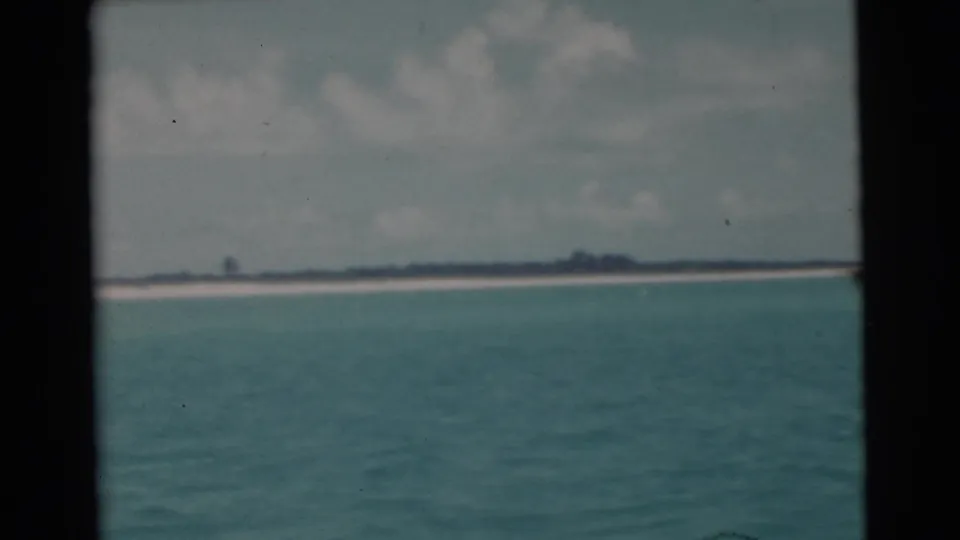 1956 Miami-area shoreline view with turquoise water in the foreground, pale beach, low vegetation, and cloudy sky.