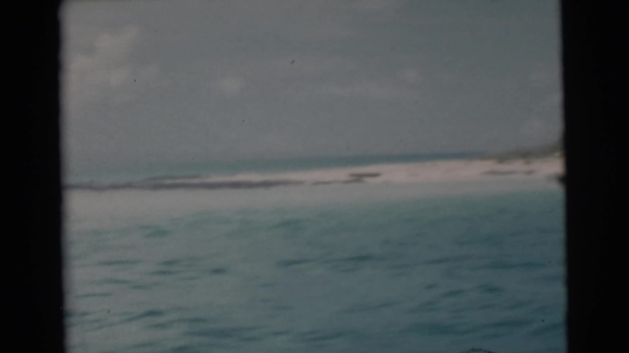 1956 Miami-area 8mm view of calm tropical water and a low beach horizon, partially bordered by the boat window or frame edge.