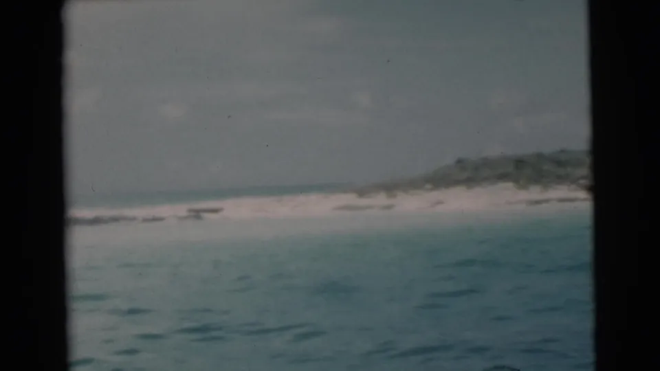 1956 Miami-area home-movie frame with blue-green water in front of a pale beach and low, dark vegetation on the horizon.