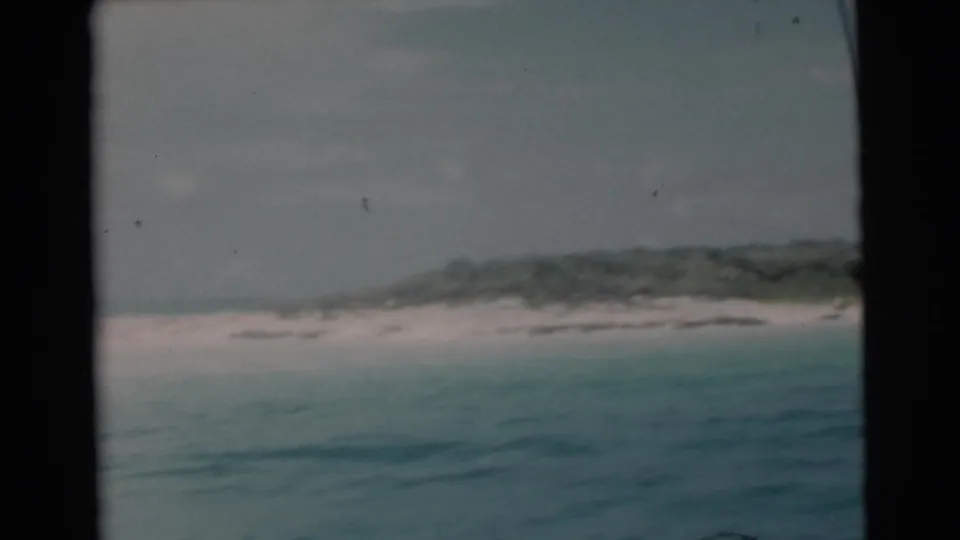 1956 Miami-area 8mm frame showing a low island-like shoreline, pale sand, and soft green-blue water under a gray sky.