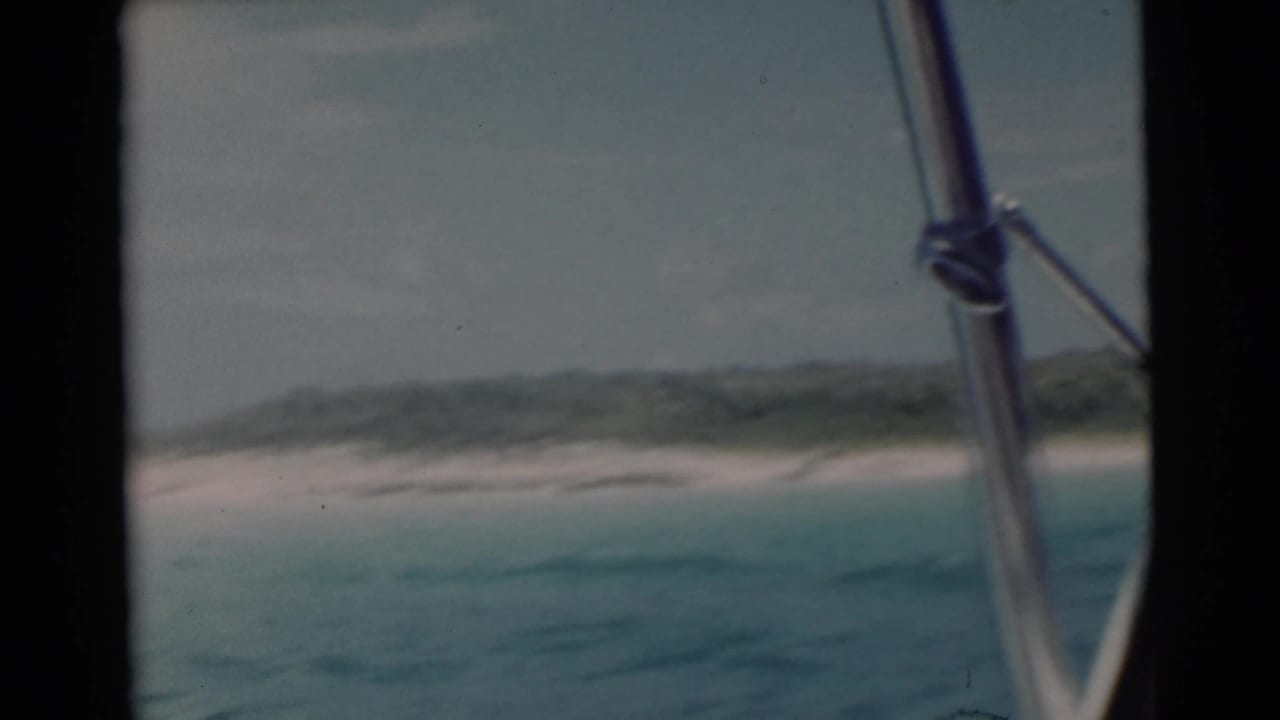 Blue water and distant sandy shore seen from a boat with metal rail in the foreground.
