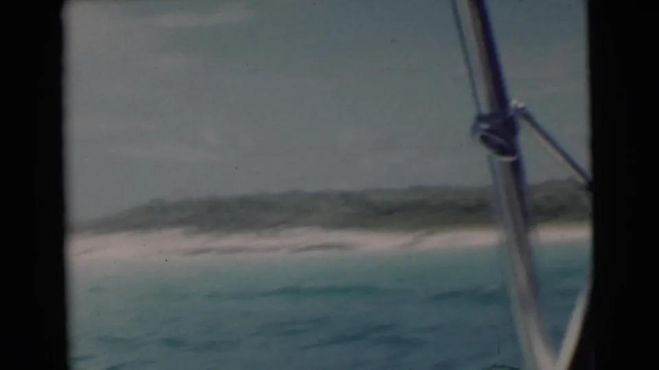 1956 Miami-area 8mm frame showing turquoise water, a pale beach, low greenery, and boat rail hardware in the foreground.