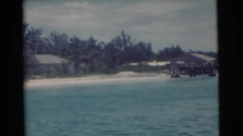 1956 8mm view near Miami showing turquoise water, sandy shore, palm trees, low buildings, and a wooden pier extending from the beach.