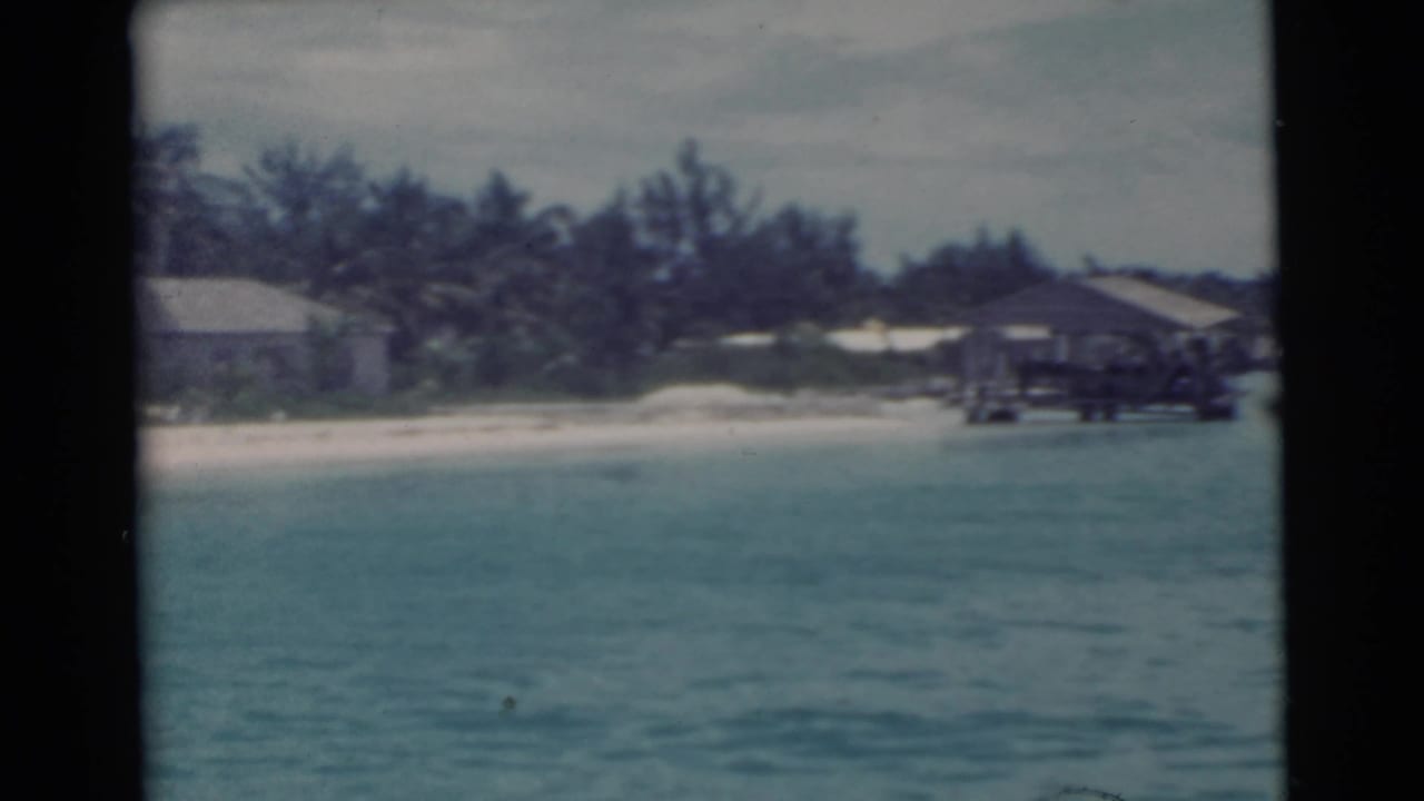 Turquoise water facing a sandy shore with low buildings and a wooden pier or dock.