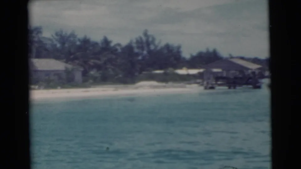 1956 Miami 8mm frame showing turquoise water, pale beach, low shoreline structures, trees, and a pier or dock from offshore.