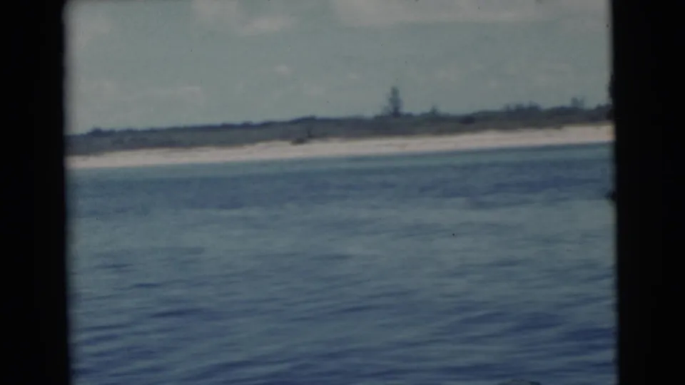 1956 Miami 8mm frame showing a long pale beach, blue water, sparse vegetation, and dark frame edges from a moving boat.