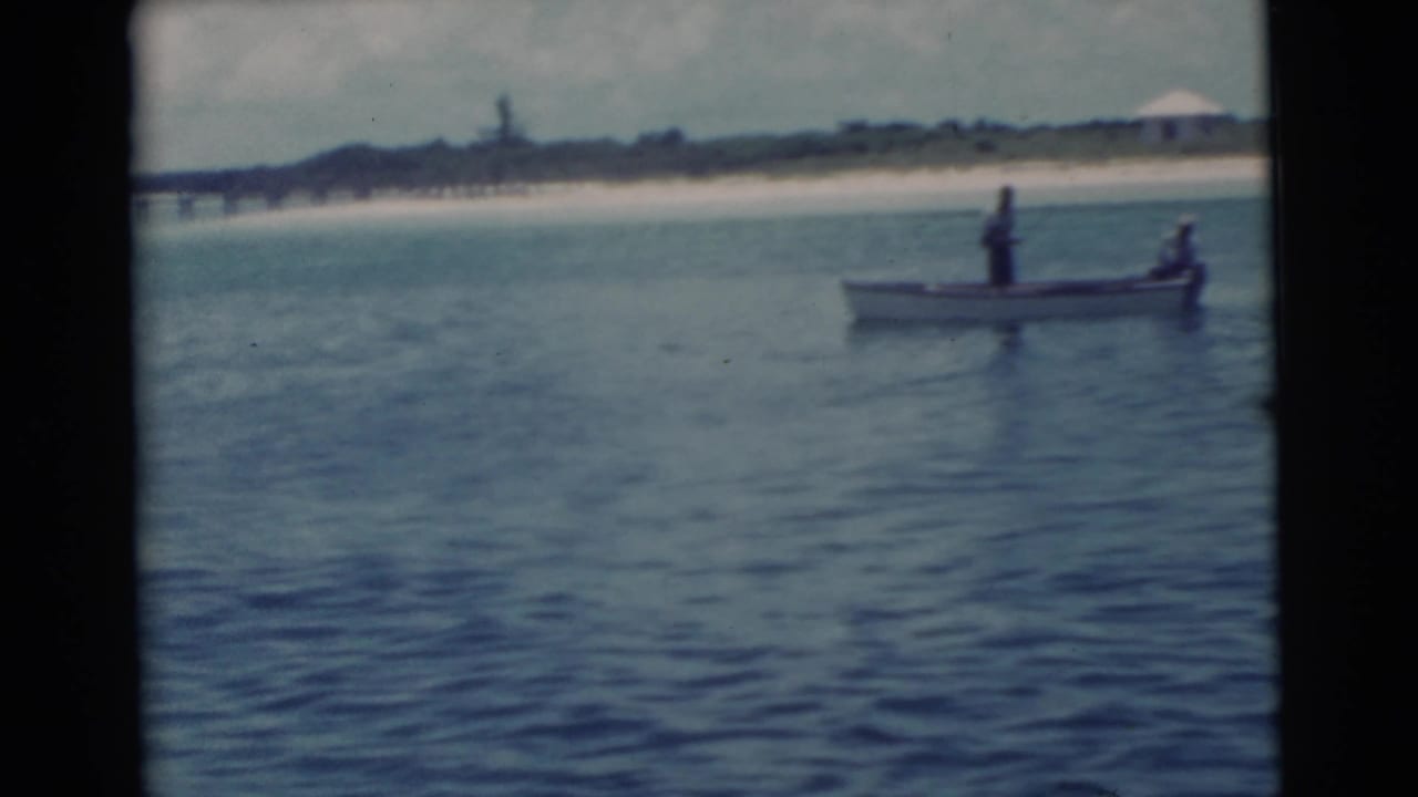 Small boat with two people on blue water near a distant beach and pier.