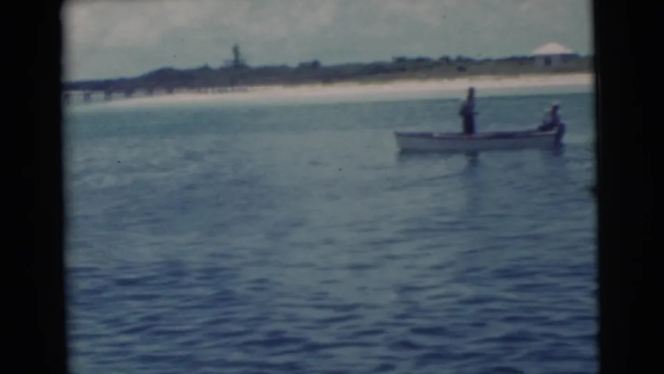 1956 Miami home-movie frame showing a small boat with two people on calm water, with a sandy shoreline and pier in the distance.