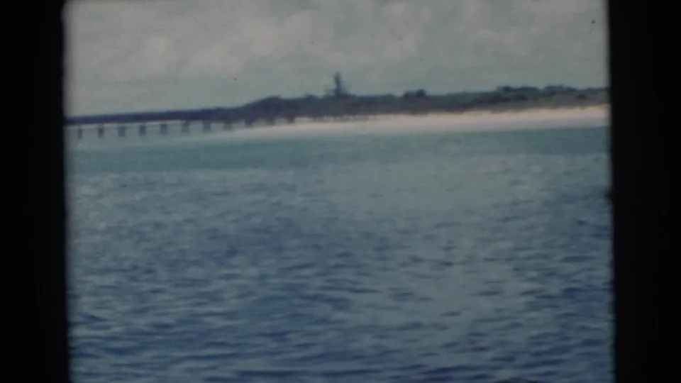 1956 Miami coastal view across blue-green water toward a long pier, sandy beach, and low shoreline under cloudy sky.