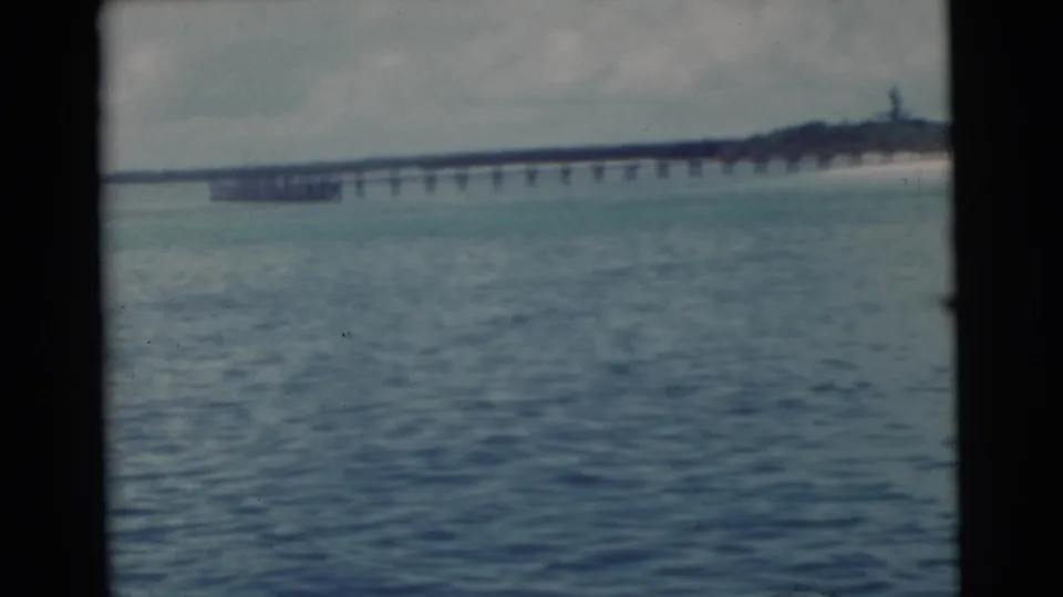 1956 Miami home-movie view of rippled water facing a long pier and sandy shoreline beneath an overcast sky.