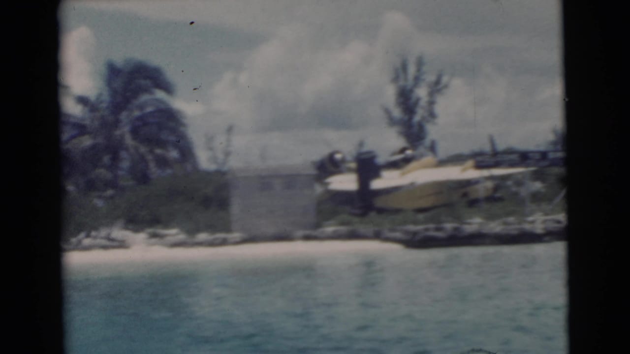 Boat-view 1956 Miami shoreline with a yellow seaplane parked near palms and rocks along the water.