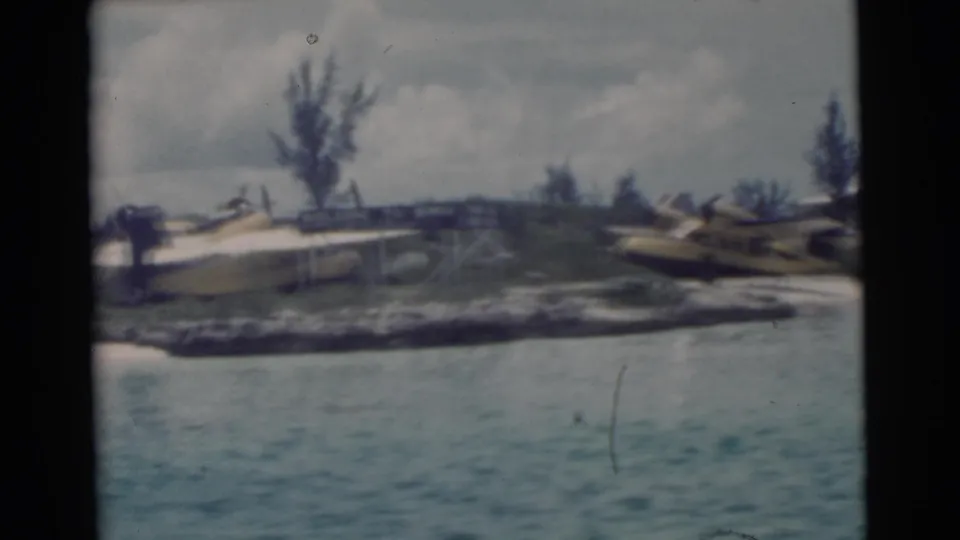 1956 Miami waterfront frame with a yellow seaplane partly centered on shore and another aircraft shape at right.