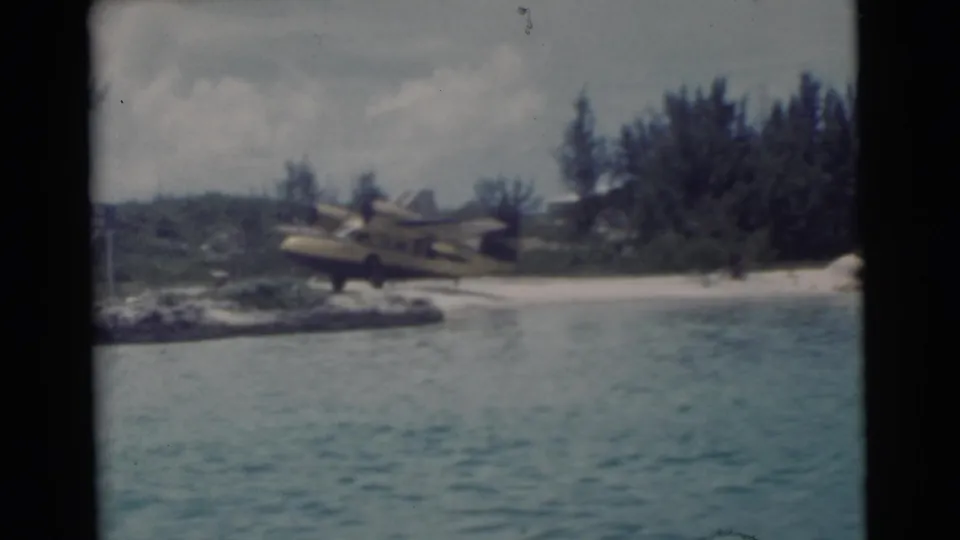 1956 Miami shoreline seen from water, with a yellow seaplane on sand, palms, and dense coastal greenery.