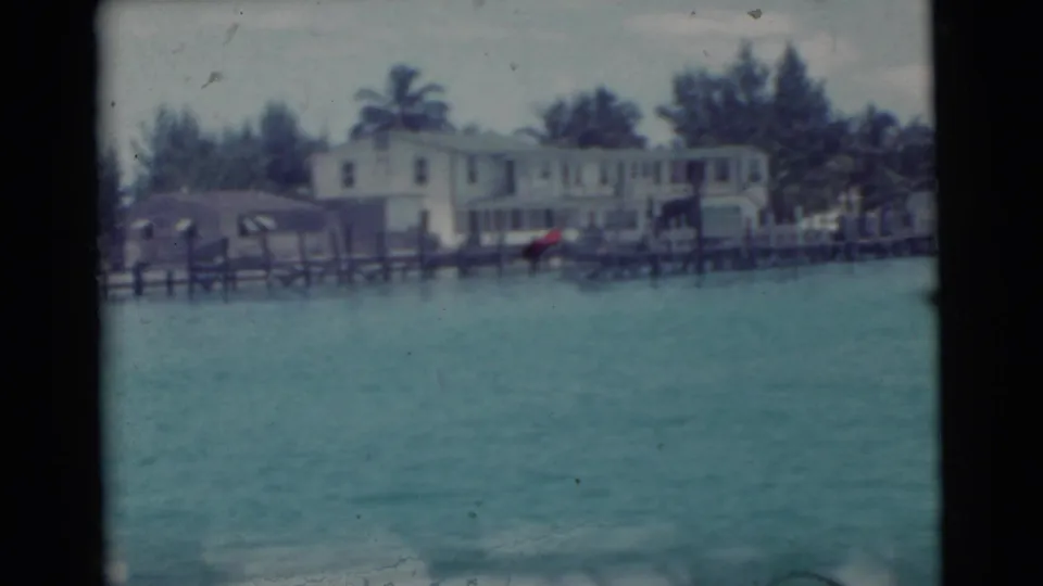 1956 Miami shoreline with waterfront residences, dock structures, palm trees, and open water in a soft 8mm frame.