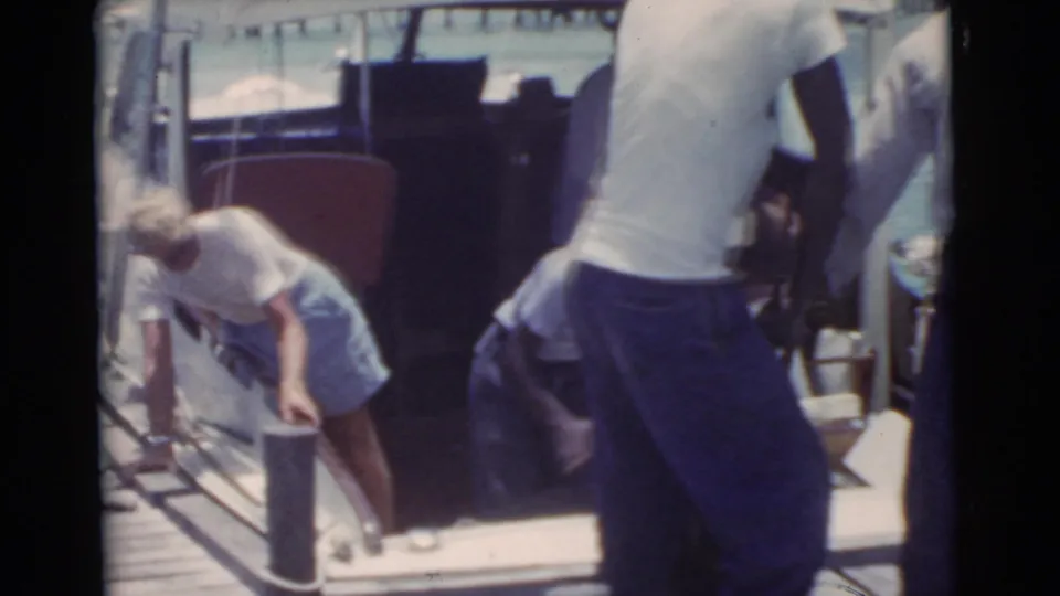 Dockside activity in 1956 Miami, with people near a moored boat and one person bending by a wooden pier post.
