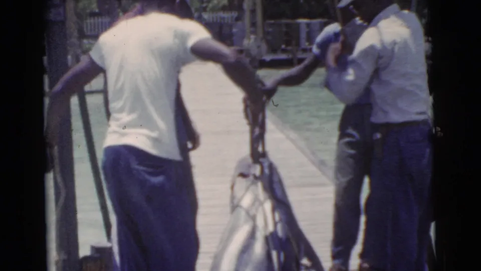 Adult men in casual shirts handle a large tuna-like fish on a Miami dock in 1956 home-movie footage.