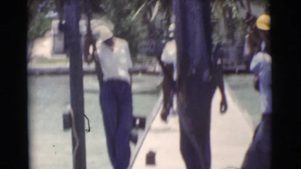 In 1956 Miami, men in light shirts and caps carry a large marlin along a waterfront dock with palms in the background.